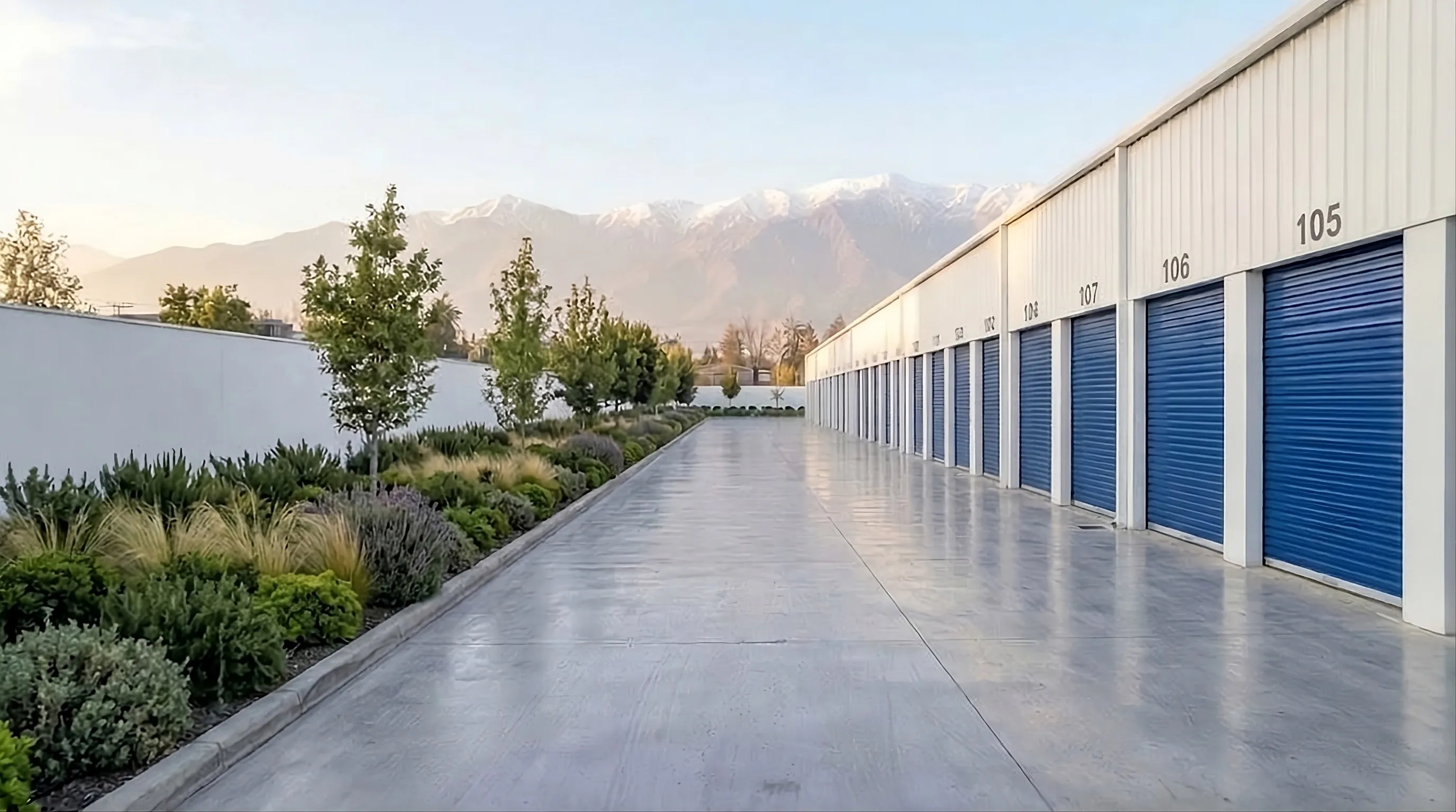 Una calle de bodegas con puertas azules numeradas, junto a un muro con jardines y árboles, y la cordillera nevada al fondo.