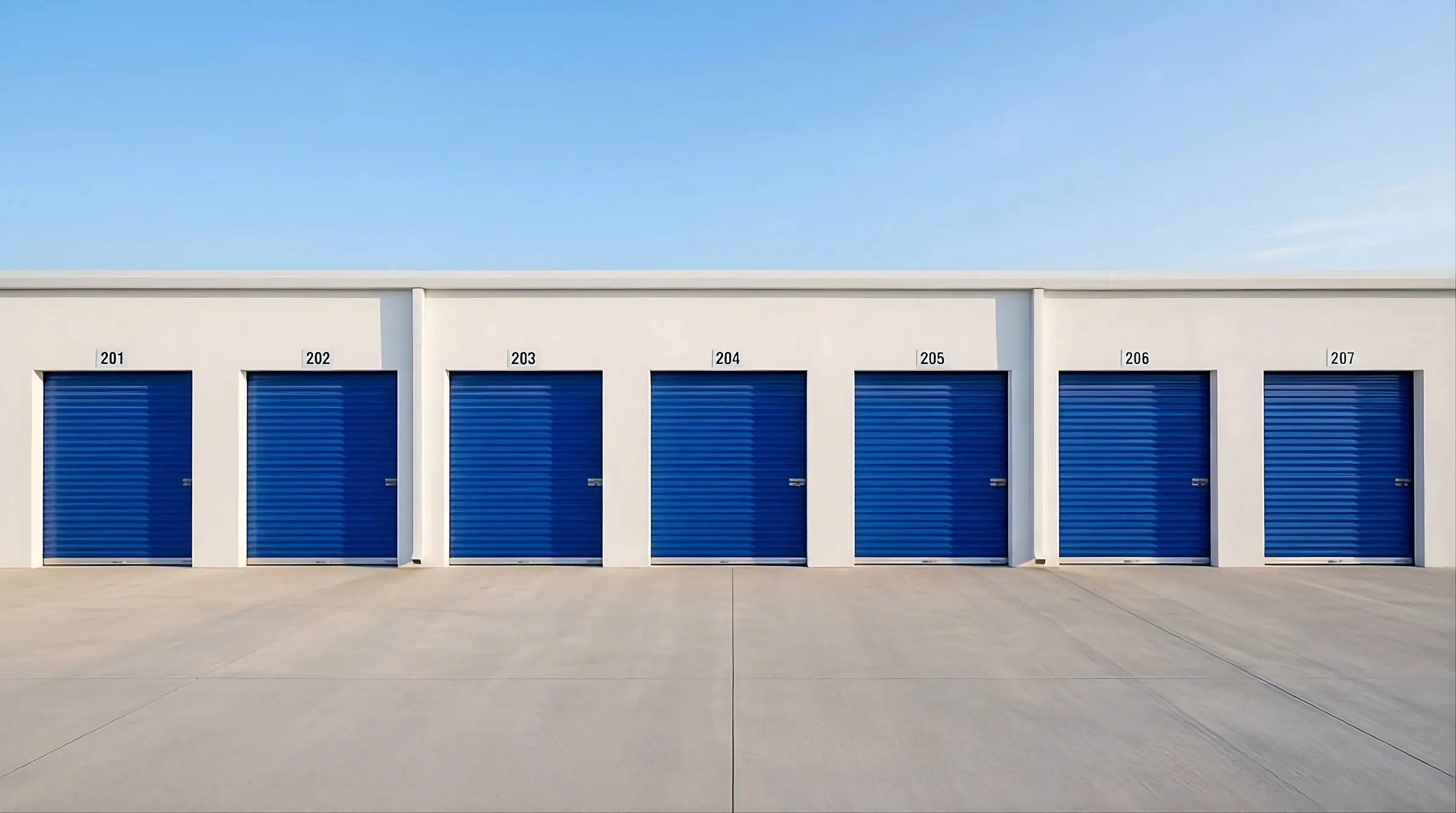 La fachada blanca de un centro de bodegas con siete puertas azules numeradas del 201 al 207, bajo un cielo despejado.