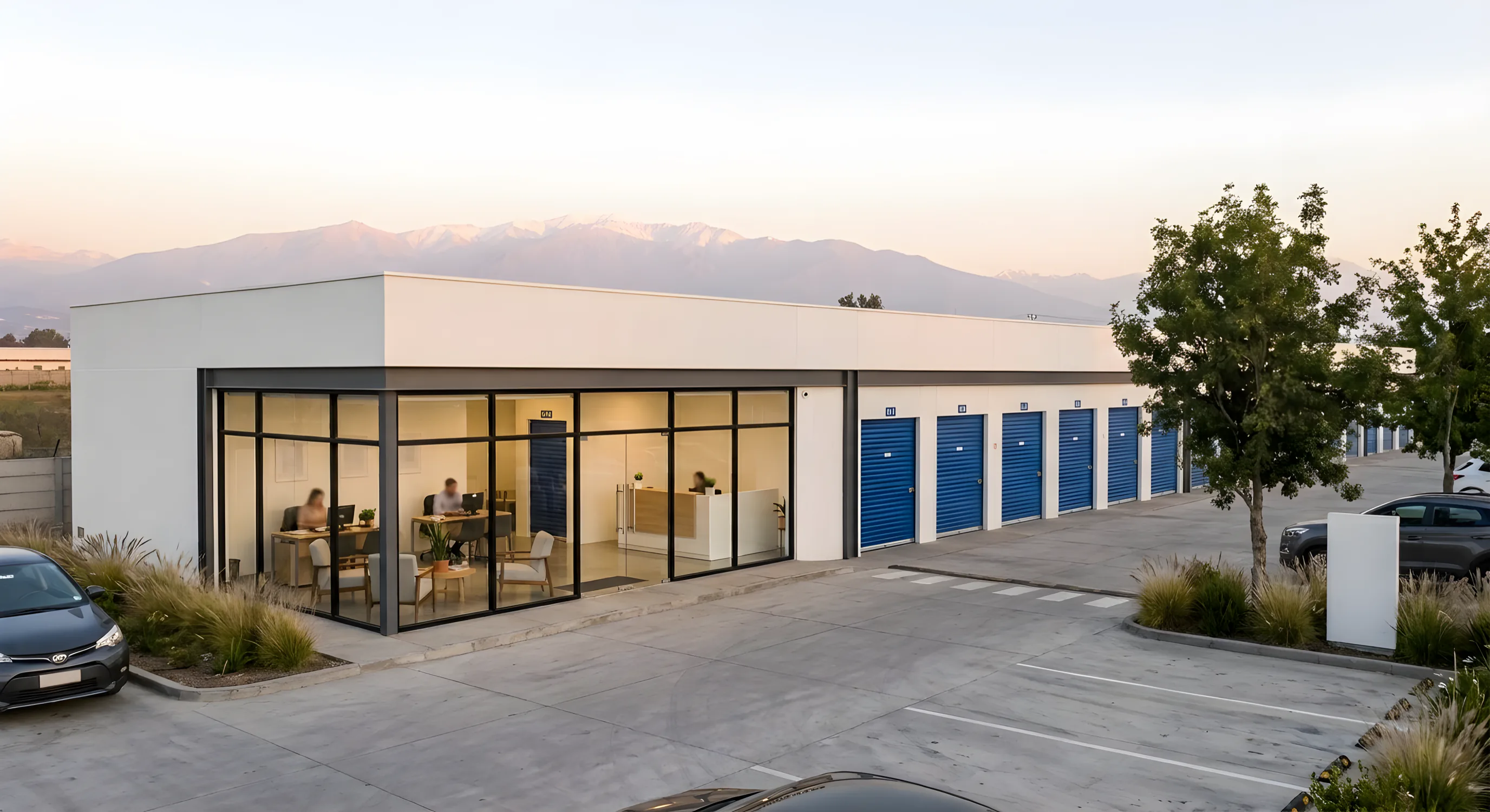 La oficina de vidrio de un centro de bodegas junto a su fila de puertas azules, al atardecer, con la cordillera de fondo.