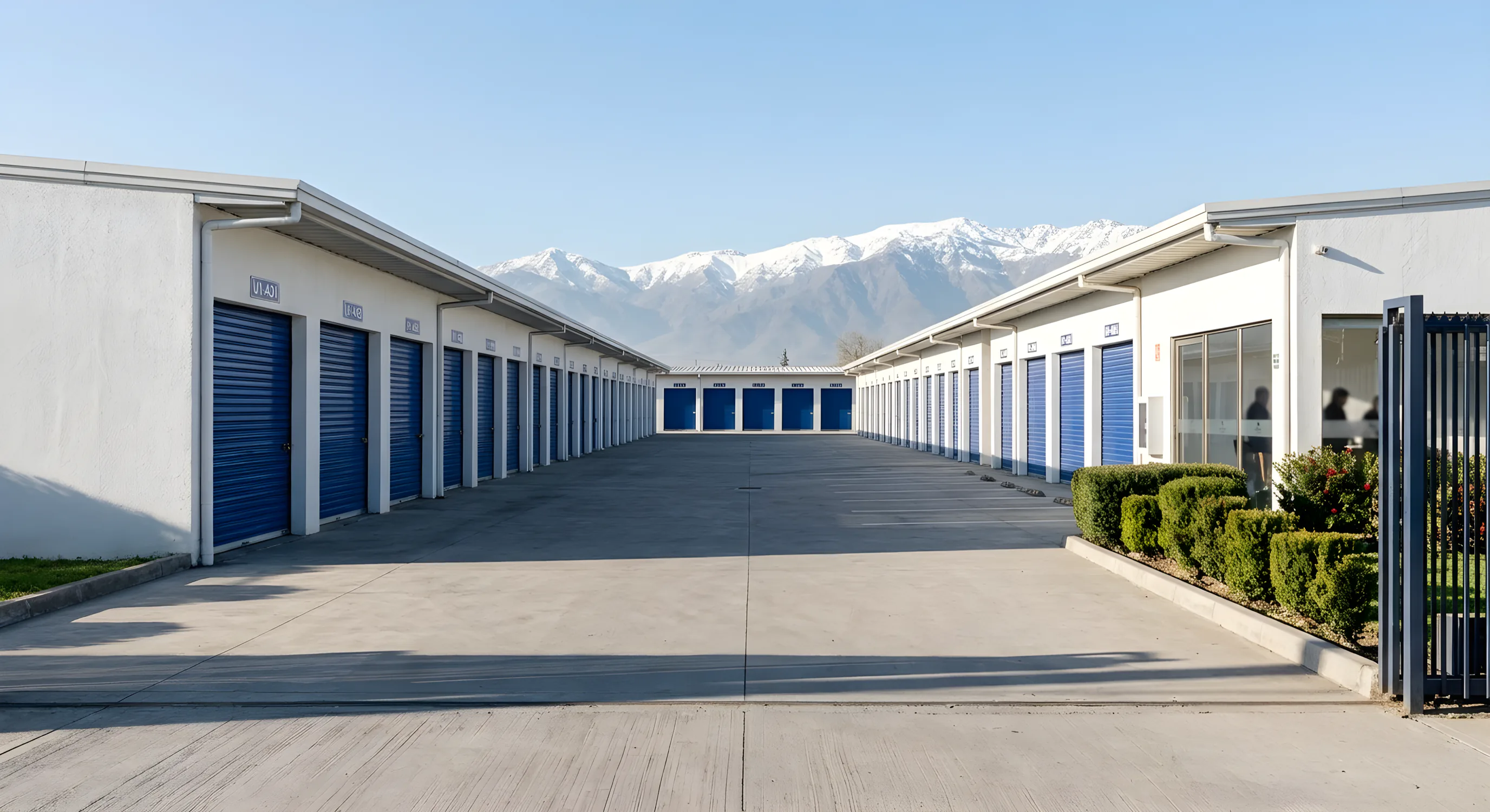 El portón abierto de un centro de bodegas, con la calle entre dos naves de puertas azules y la cordillera nevada al fondo.